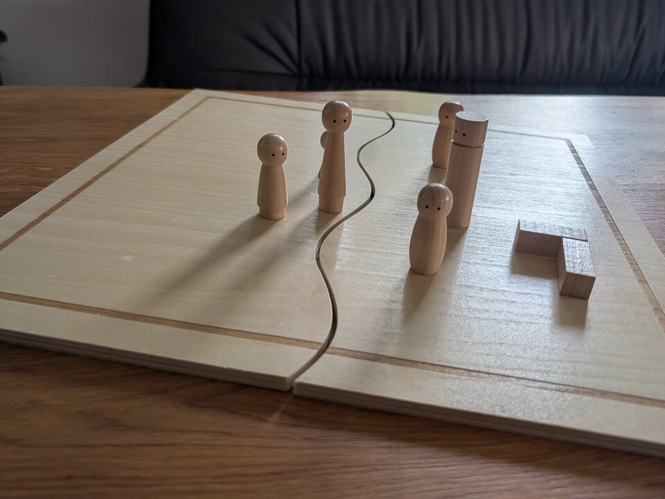 Ein Systembrett aus hellem Holz auf einem Tisch: Eine dunkle Schnur teilt die Fläche in zwei Hälften, links stehen zwei Holzfiguren, rechts zwei weitere und daneben zwei kleine Holzklötze.